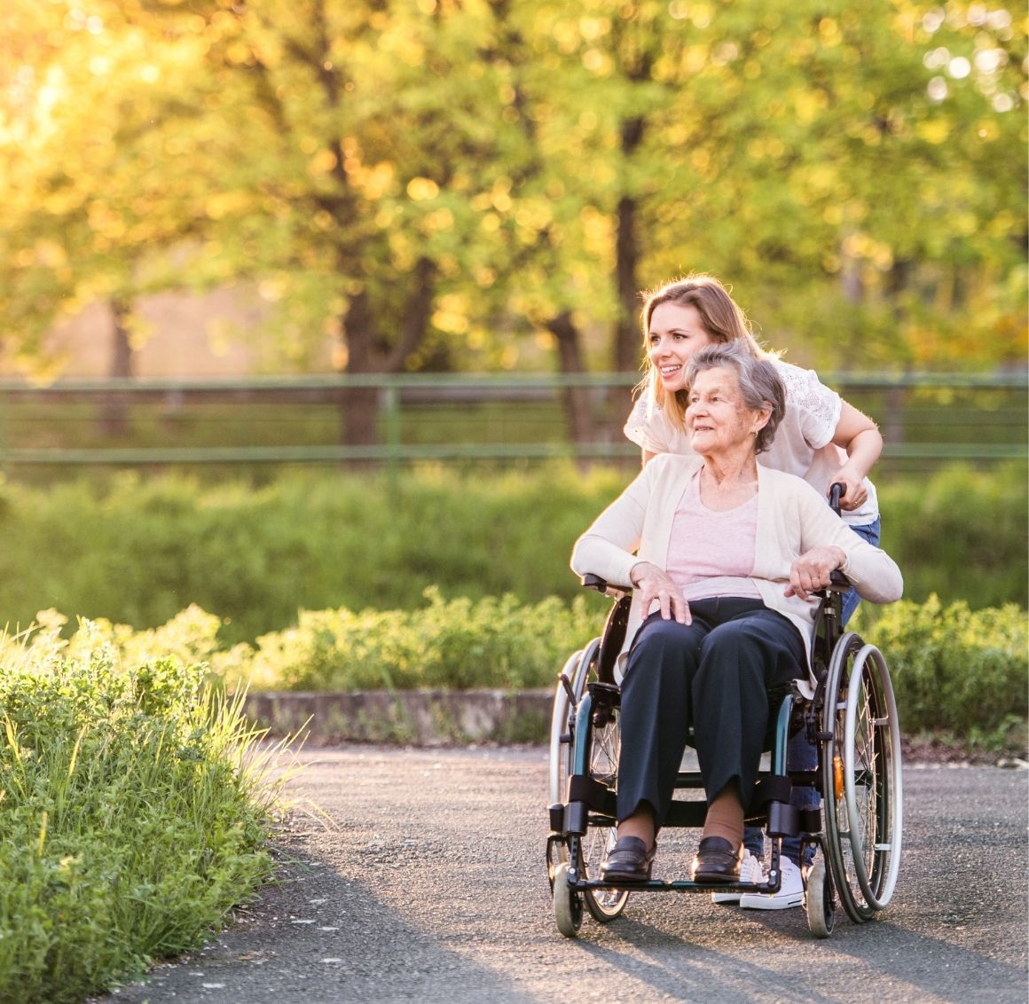 A young woman caregiver pushing and elderly woman in a wheel chair outside on a path.