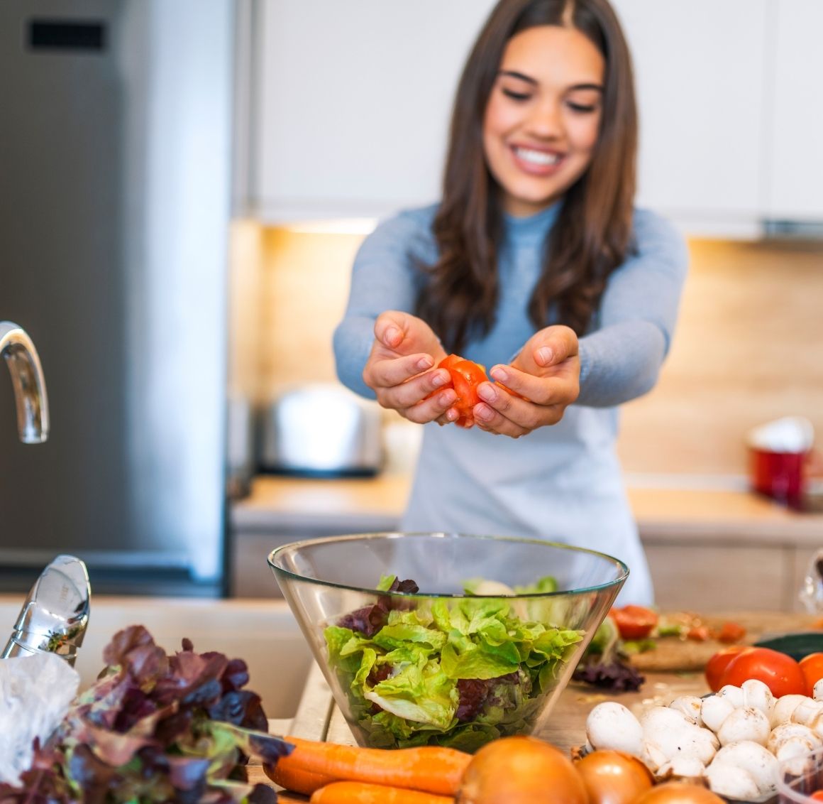 A young woman with dark hair in a kitchen with a hand full of tomatoes over a bowl of salad.