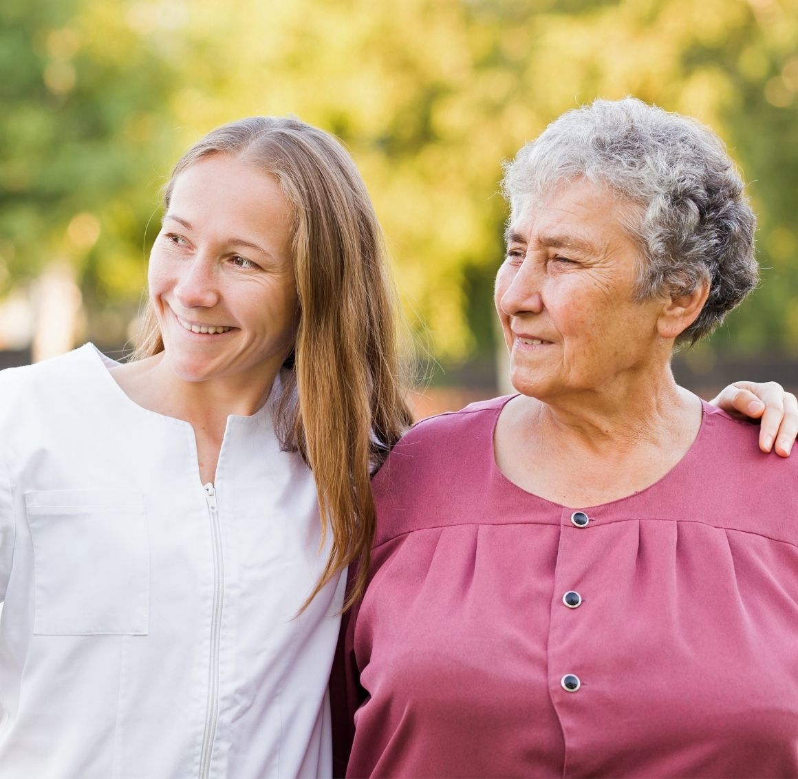 A woman with her arm around and elderly woman, walk out side looking in the distance.