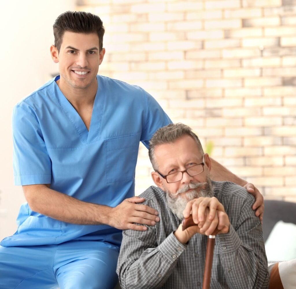 An older man sitting with hands one cane, accompanied by male caregiver in blue with hand on elderly persons shoulder.