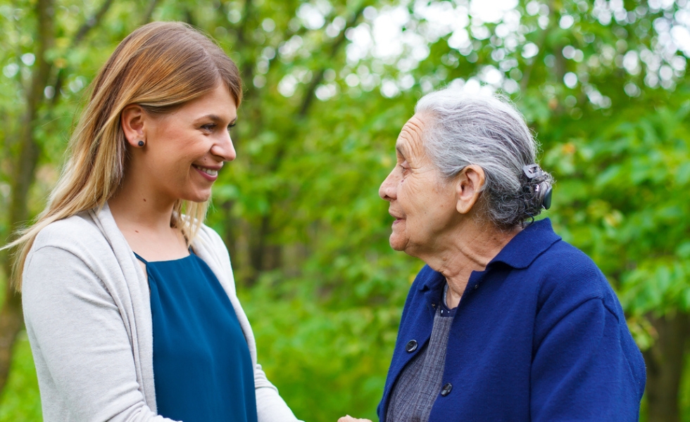 Outdoors and surrounded by greenery, a young girl with long hair holds hands with a smiling senior woman as they gaze at each other, sharing a joyful and heartfelt moment.