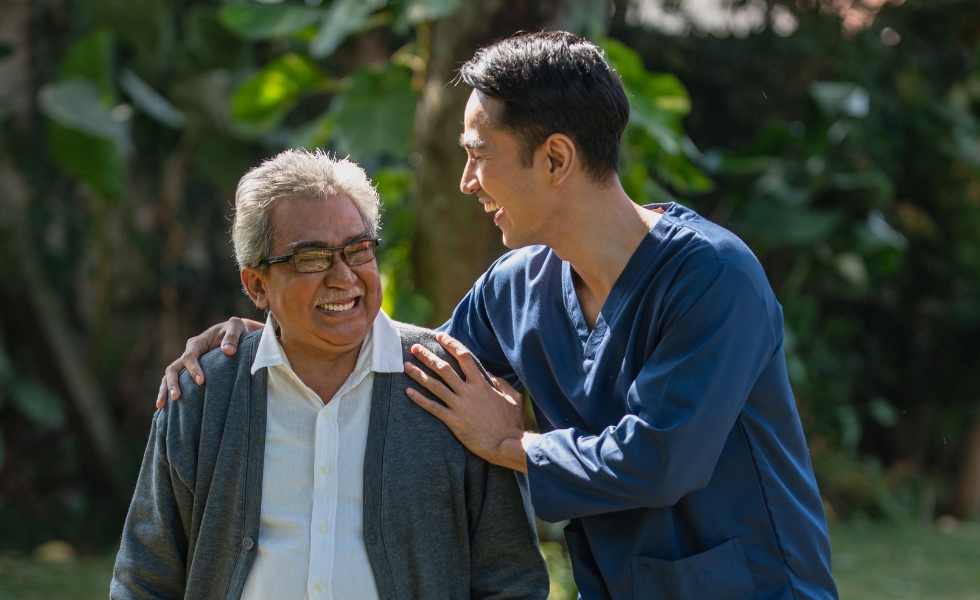 A cheerful male caregiver laughs and holds shoulder of elderly man as they walk in a park. Companionship Care in Chicago