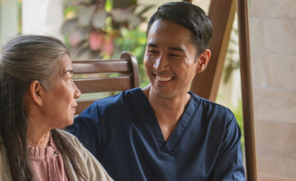 Personal Care for Seniors A smiling male caregiver in blue scrubs provides personal care in Columbia, sitting beside a senior woman and engaging in a warm conversation outdoors.
